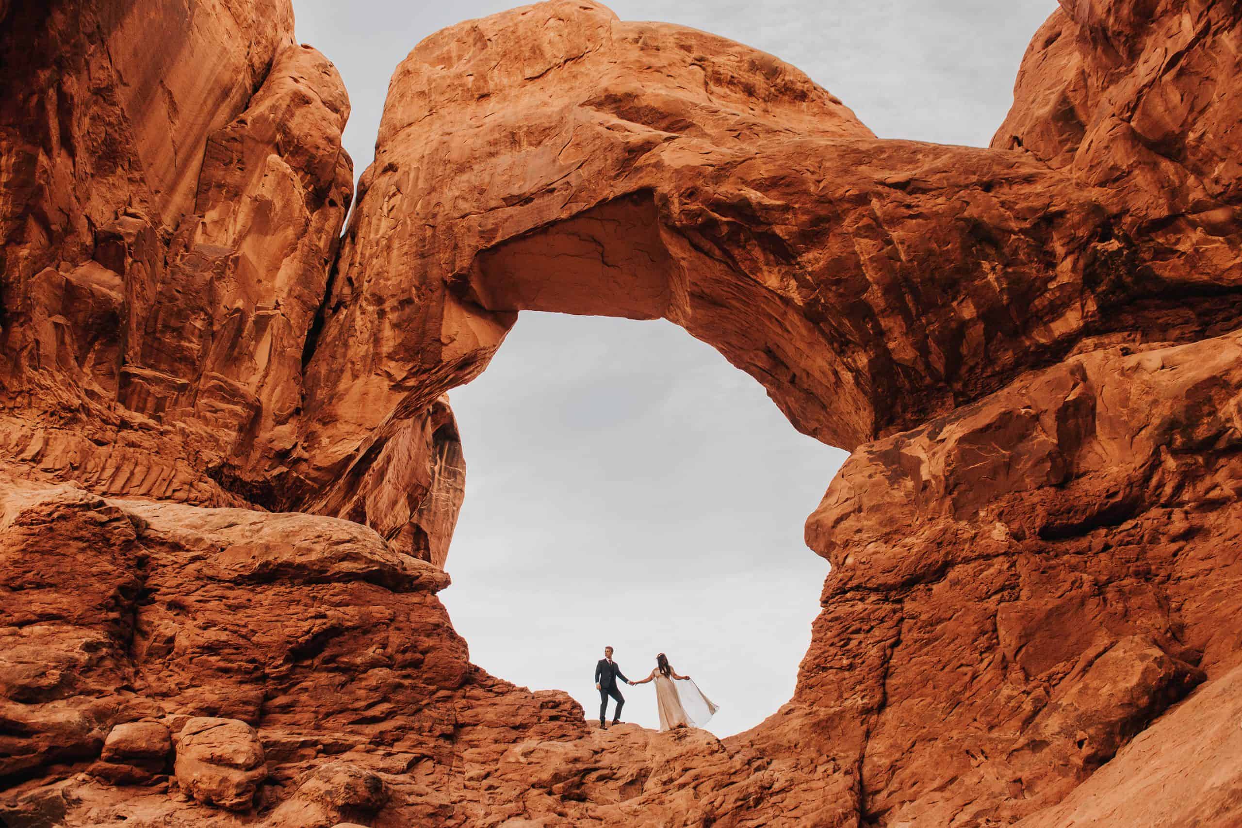 adventure bride and groom holds hands far away while standing under an arch in Arches National Park
