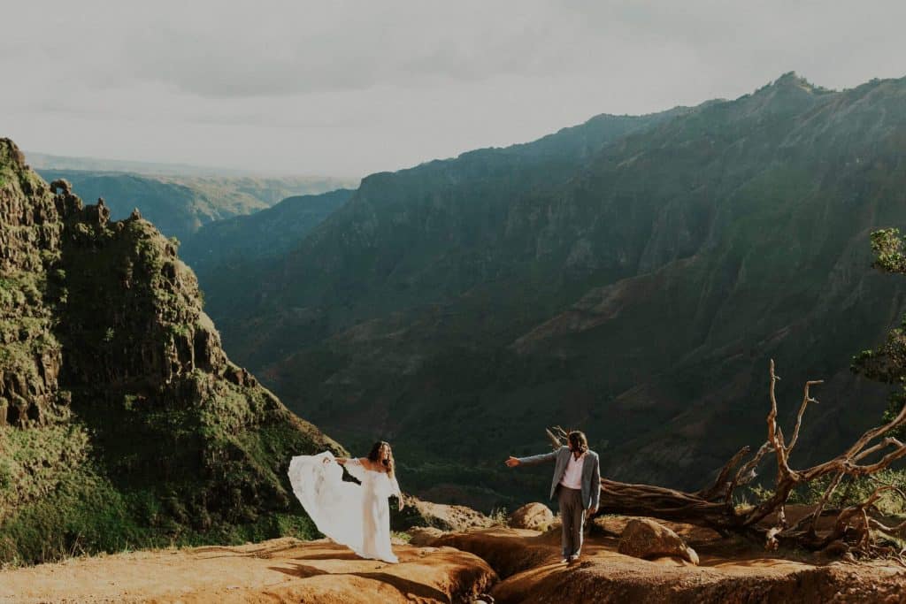 bride and groom reach for each other while standing on a cliffside in Waimea Canyon in Kauai, Hawaii