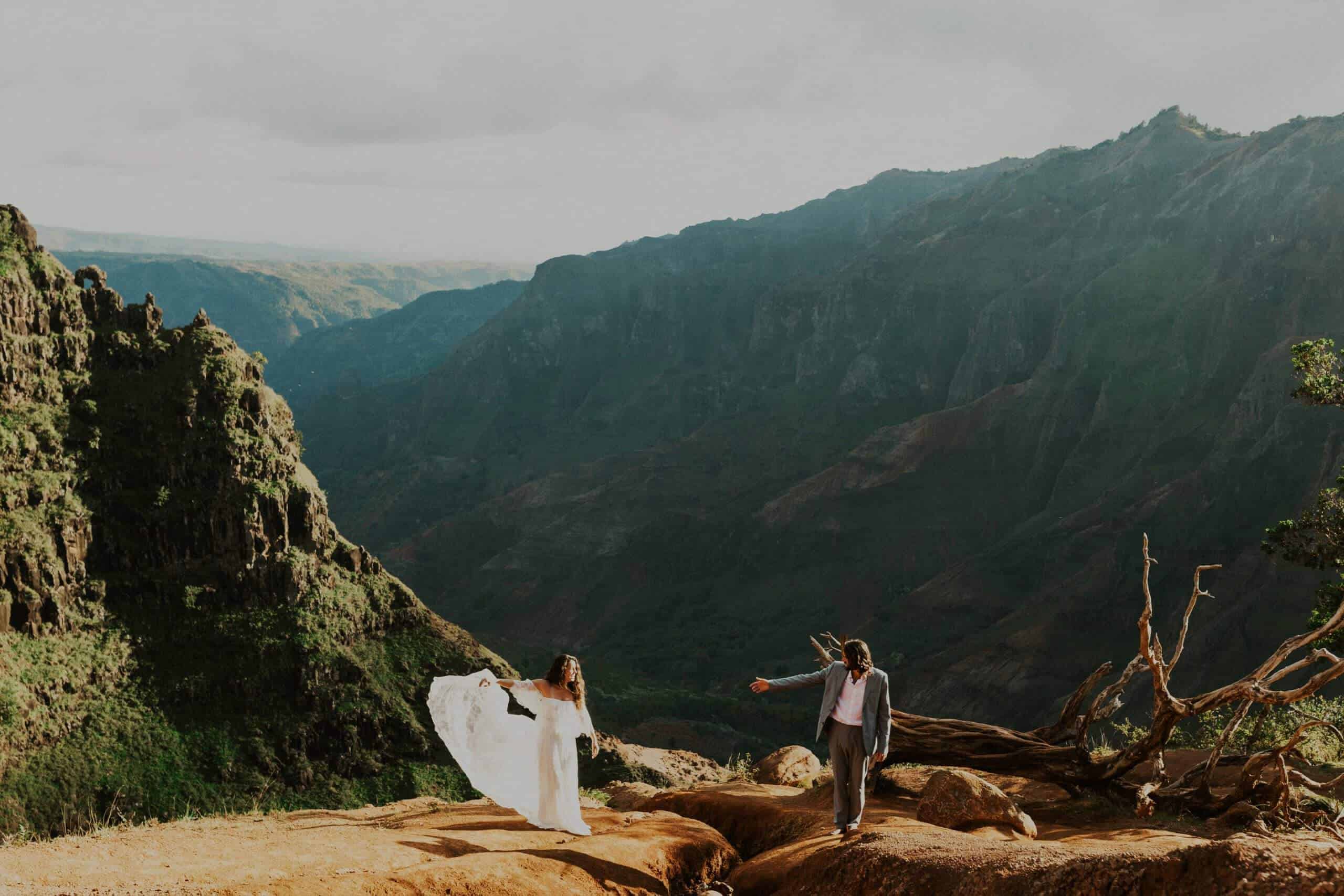 adventure elopement bride and groom reach for each other while standing on a cliffside in Waimea Canyon in Kauai, Hawaii